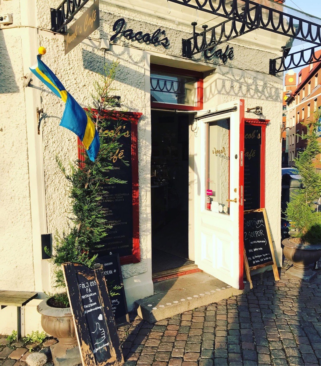 Entrén till Jacobs Café med öppen dörr, svensk flagga och tavlor på trottoaren